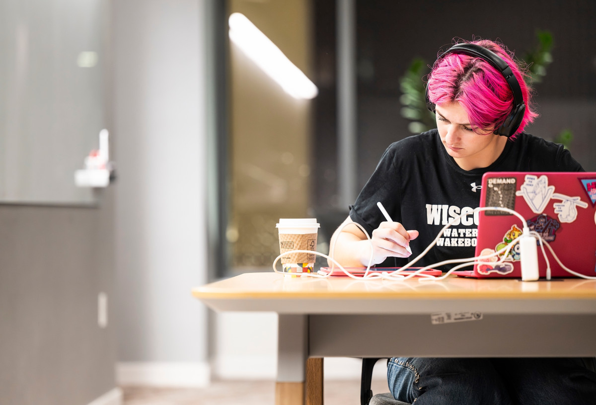 A student sits alone at a table writing notes on a tablet while wearing headphones.