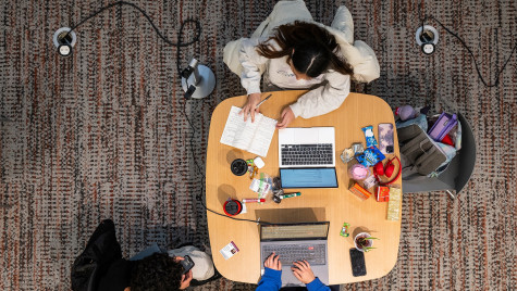 From above, three students sit at a table reviewing notes and working on their laptops.