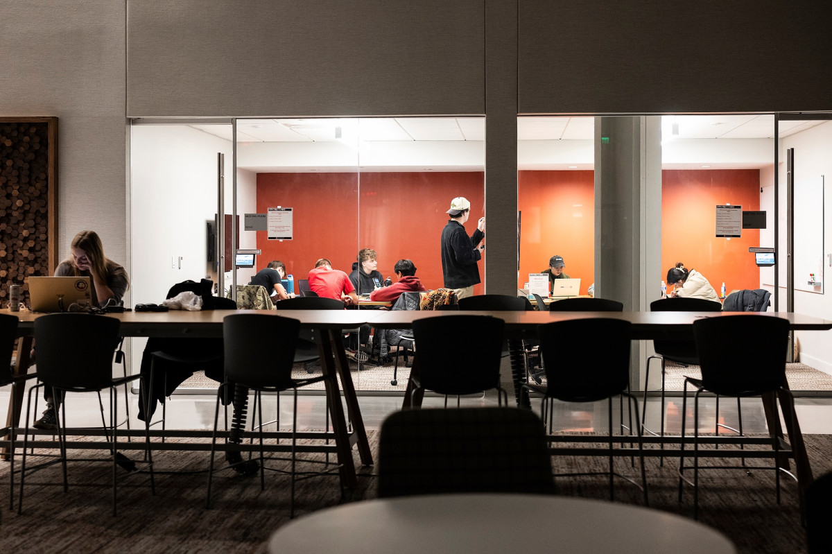 From the outside of a glass study room, a group of students sit around a table and collaborate on studying.