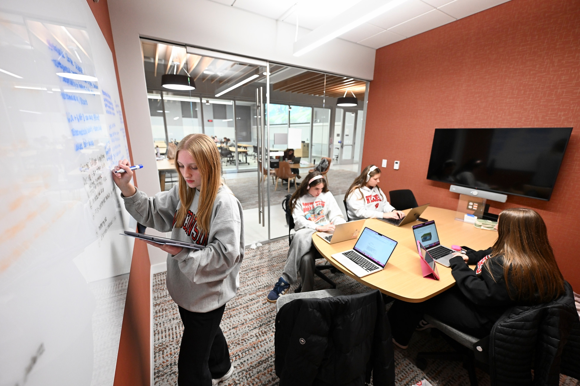 In a small study room, a group of students sit around a table, while one stands at a white board on the wall and transcribes notes.