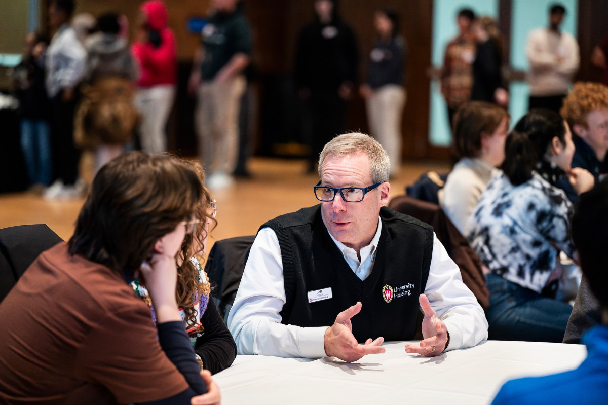 Novak sits next to two students at a table and engages in conversation with while dinner is served.