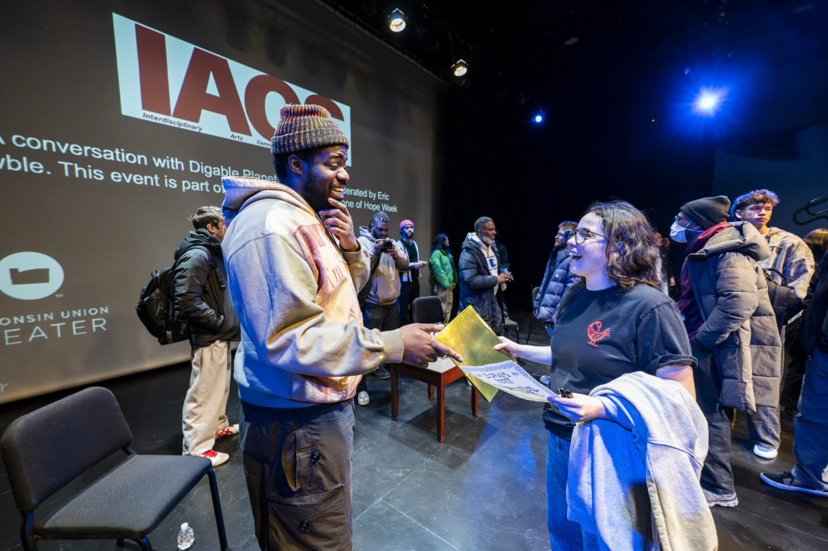 A man stands on stage and talks to a young woman who holds an event flier in her hand.