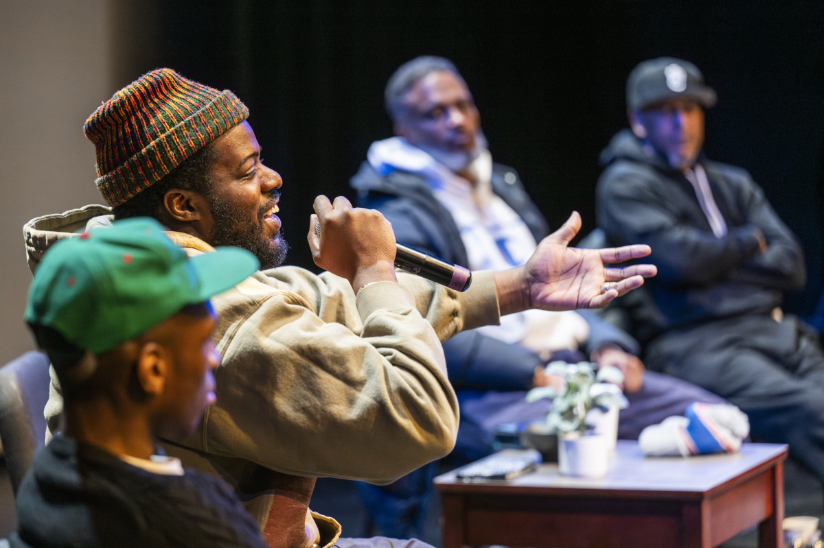 MIKE sits on stage with a microphone in his hand and speaks to the audience.
