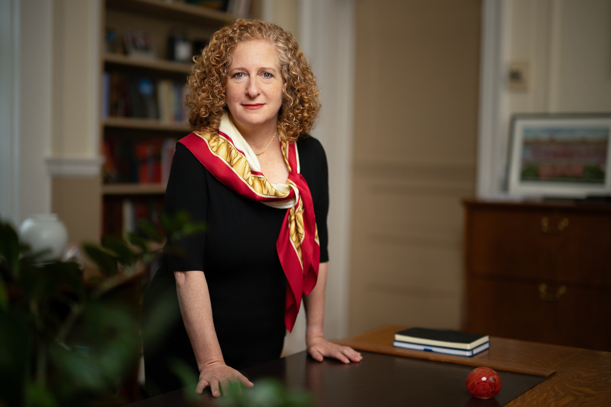 A woman wearing a black dress and a red, gold and white scarf stands at a desk, looking into the camera. She has red, curly hair and red lipstick.