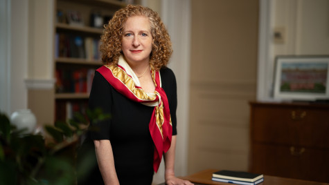 A woman wearing a black dress and a red, gold and white scarf stands at a desk, looking into the camera. She has red, curly hair and red lipstick.