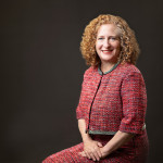Chancellor Jennifer Mnookin is seated against a dark background wearing a textured red jacket and skirt with black-and-white trim and their hands folded in their lap.