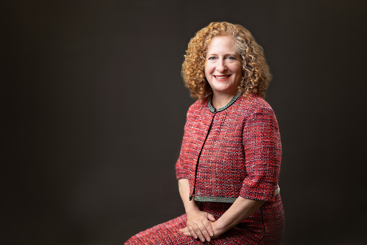 Chancellor Jennifer Mnookin is seated against a dark background wearing a textured red jacket and skirt with black-and-white trim and their hands folded in their lap.