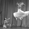 In a black and white photo, Woodbury is pictured suspended in air in mid-dance move. A woman sits on the stage and plays a drum with her hands during the dance.