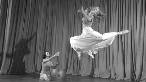 In a black and white photo, Woodbury is pictured suspended in air in mid-dance move. A woman sits on the stage and plays a drum with her hands during the dance.