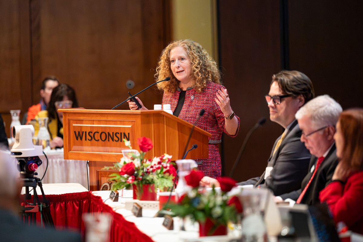 A woman stands at a podium and talks and gesticulates as others listen.