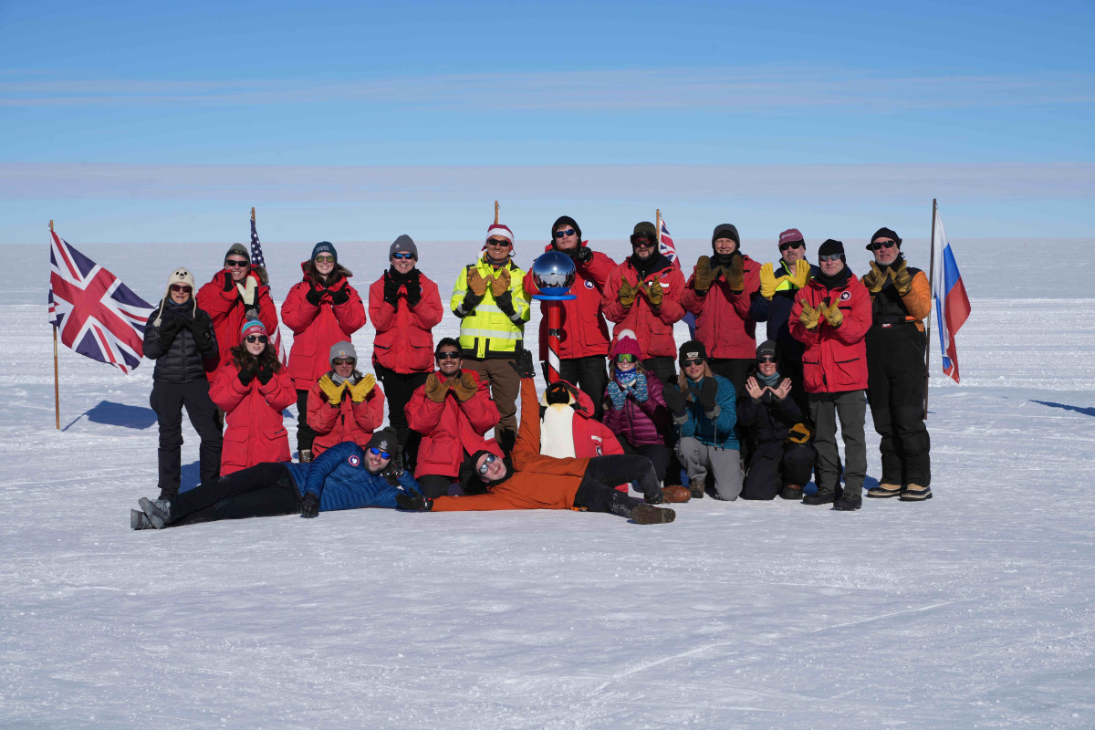 A group of researchers wearing heaving layers of winter clothes stand outside and pose for a photo. Many are holding up their hands in the W pattern.