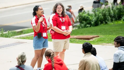 Two UW-Madison students, both wearing red tour guide shirts, stand in front of prospective students who are seated on the grass. One tour guide holds a small microphone while addressing the group.