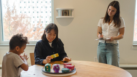 Pruchnicki stands with a clipboard in her hand while a mother and child sit at a table and play with brightly colored blocks.