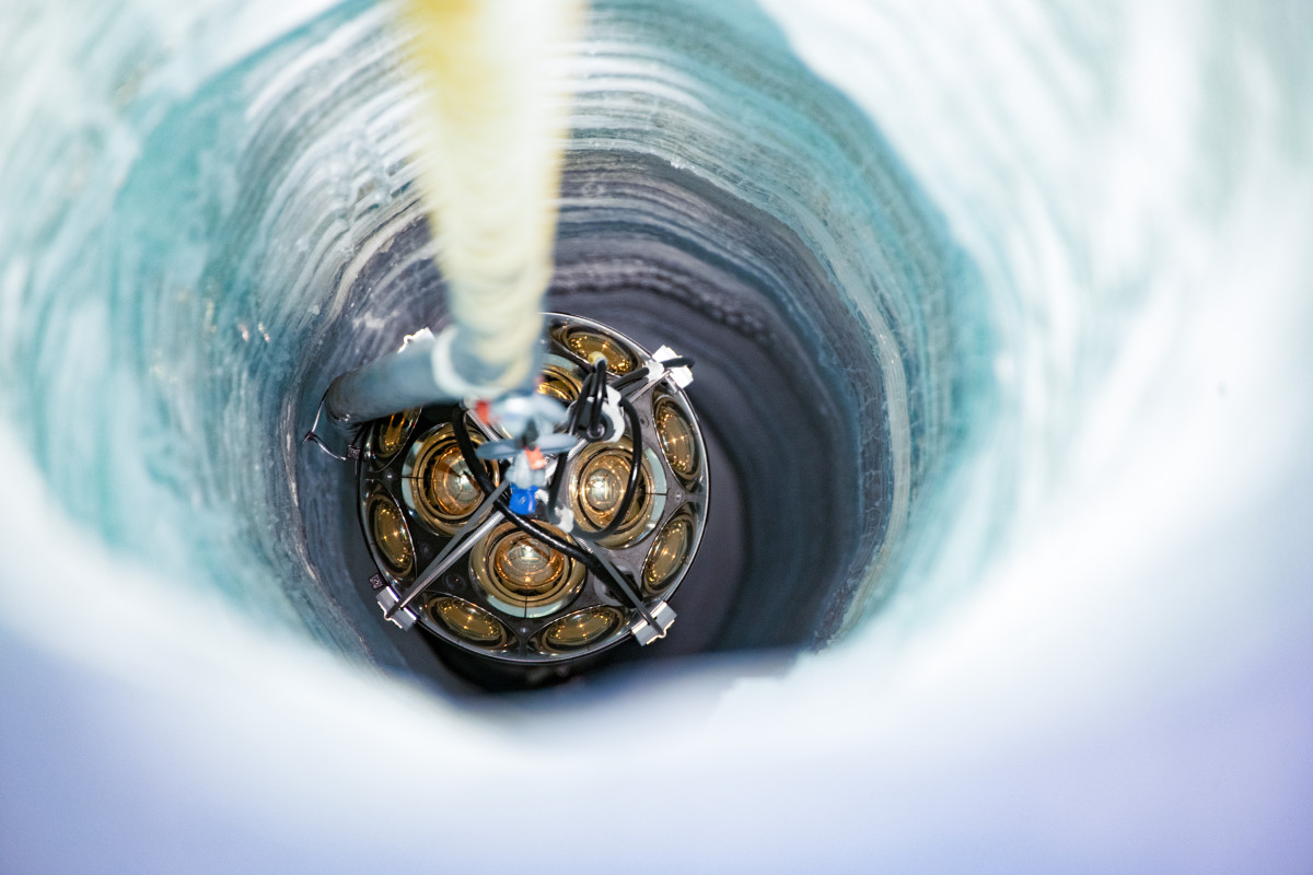 A photo shows an overhead view of a glass orb being lowered into a drilled hole in thick layers of ice.
