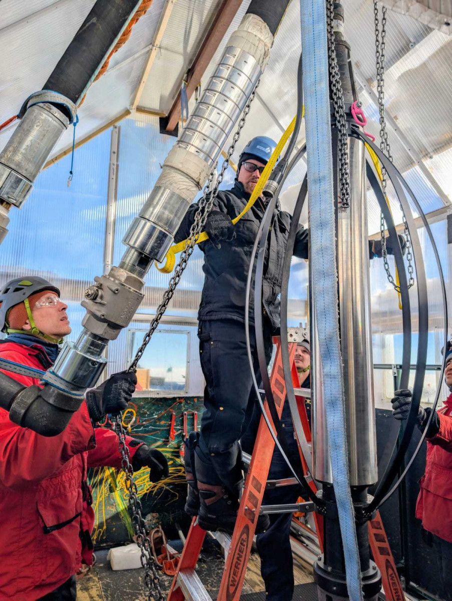 Four people surround a massive metal drill and prepare to push it down into the ice.