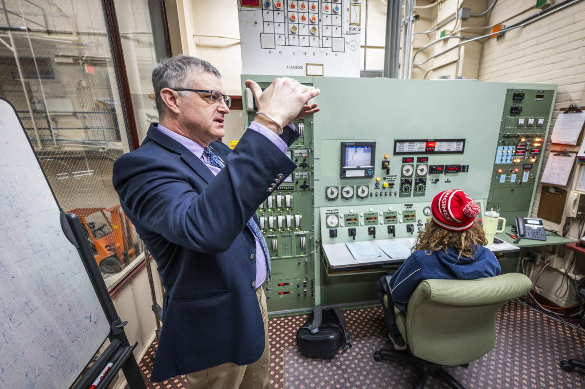 Wilson explains the mechanics of the nuclear reactor, using his hands to demonstrate the concepts. He stands behind a graduate student who operates the control panel.