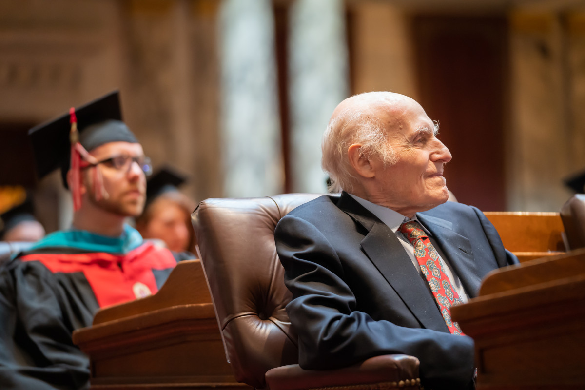 In the foreground, Senator Kohl sits in a brown leather chair. Behind him sits a group of graduates wearing caps and gowns.