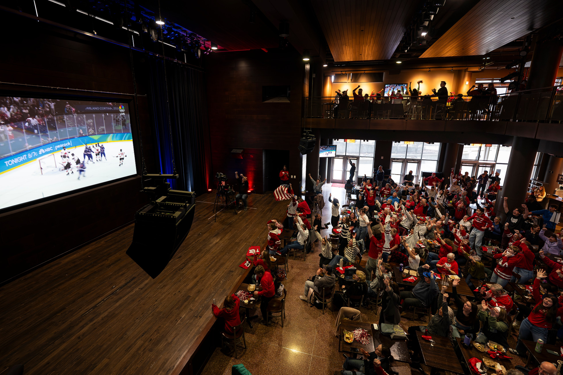 A photo taken from above shows Badger fans wearing red jerseys filling the open space of The Sett below as they watch a large screen projecting a hockey game.