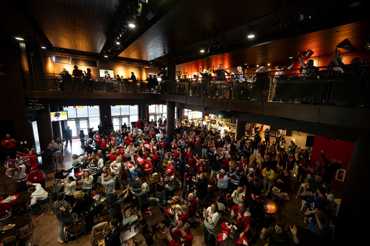 A photo taken from above shows Badger fans wearing red jerseys filling the open space of The Sett below as they watch a large screen projecting a hockey game.