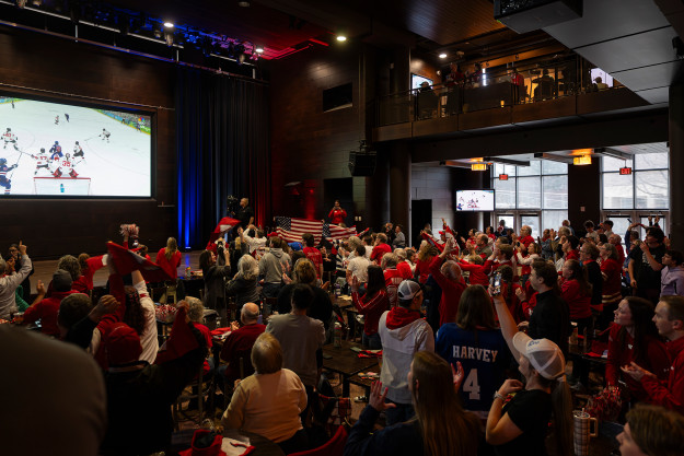 Badger fans wearing red jerseys and holding American flags fill the open space of The Sett as they watch a large screen projecting a hockey game.