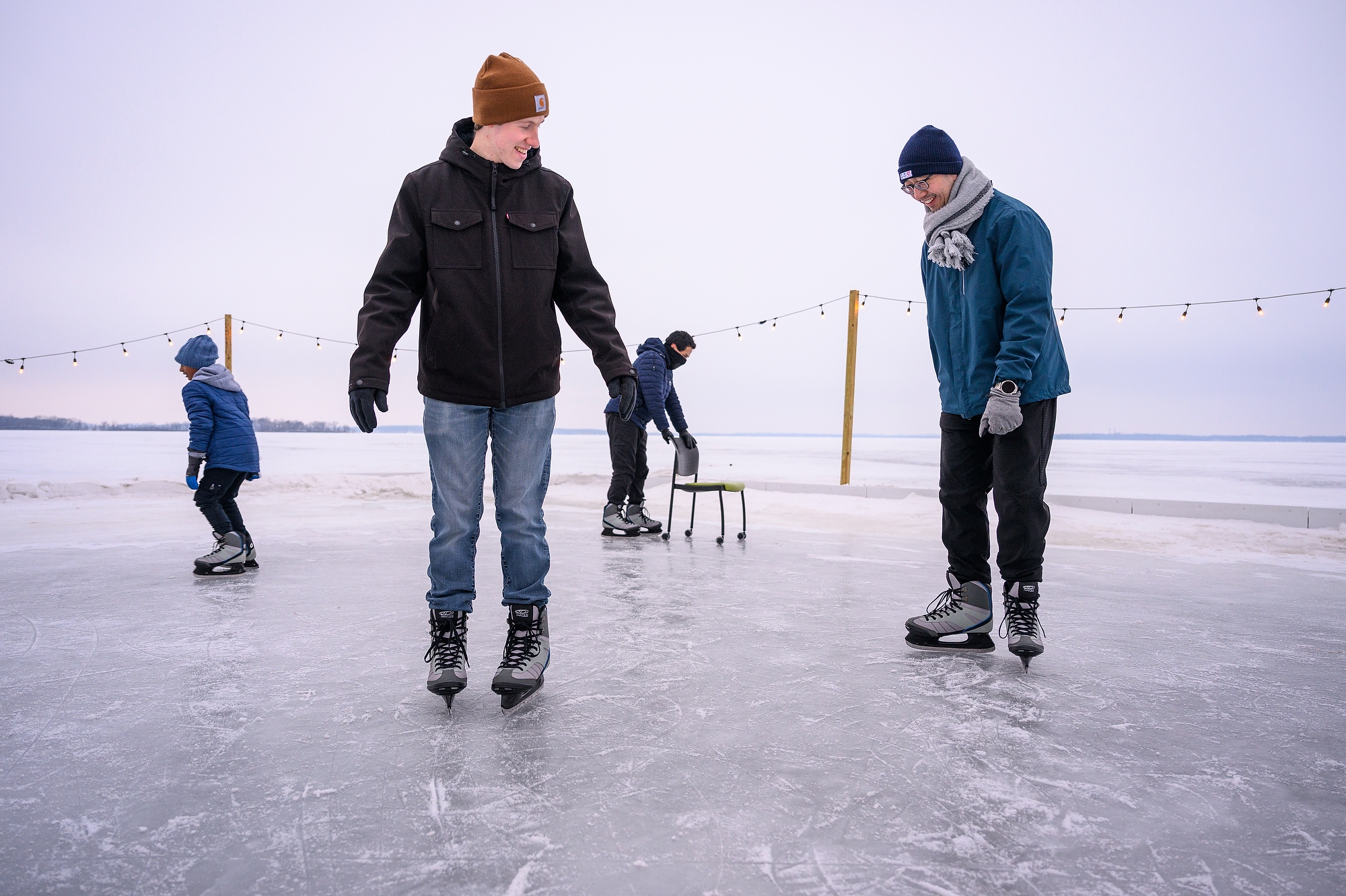 A man demonstrates an ice skating move as his friends watches. In the background, people skate and move chairs on the ice.
