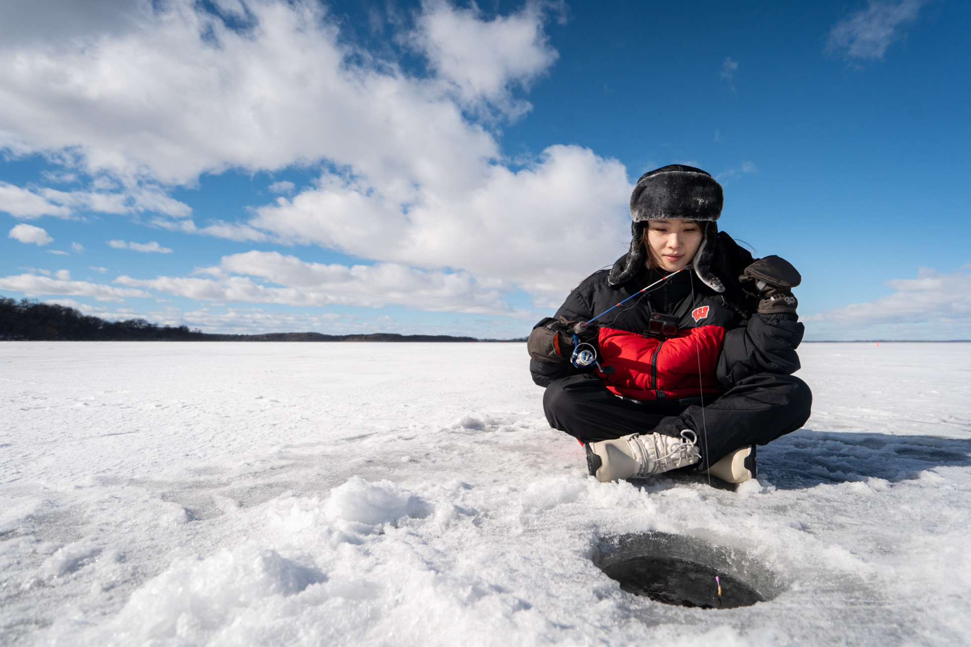 A woman learning how to ice fish sits on the ice holding a fishing rod and reel over a hole drilled in front of her.