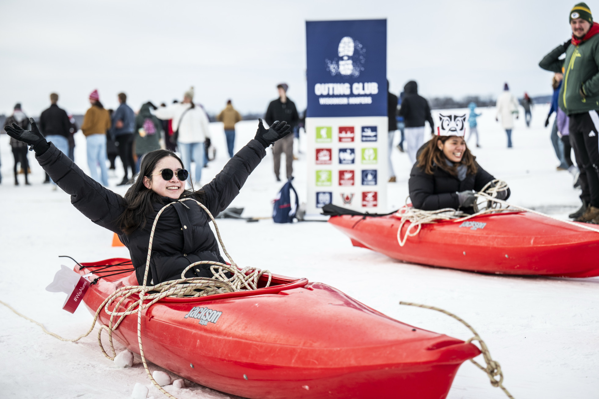 Two women are sitting in red kayaks on the ice. One holds her arms up and smiles in triumph after winning the kayak ice pulley race.