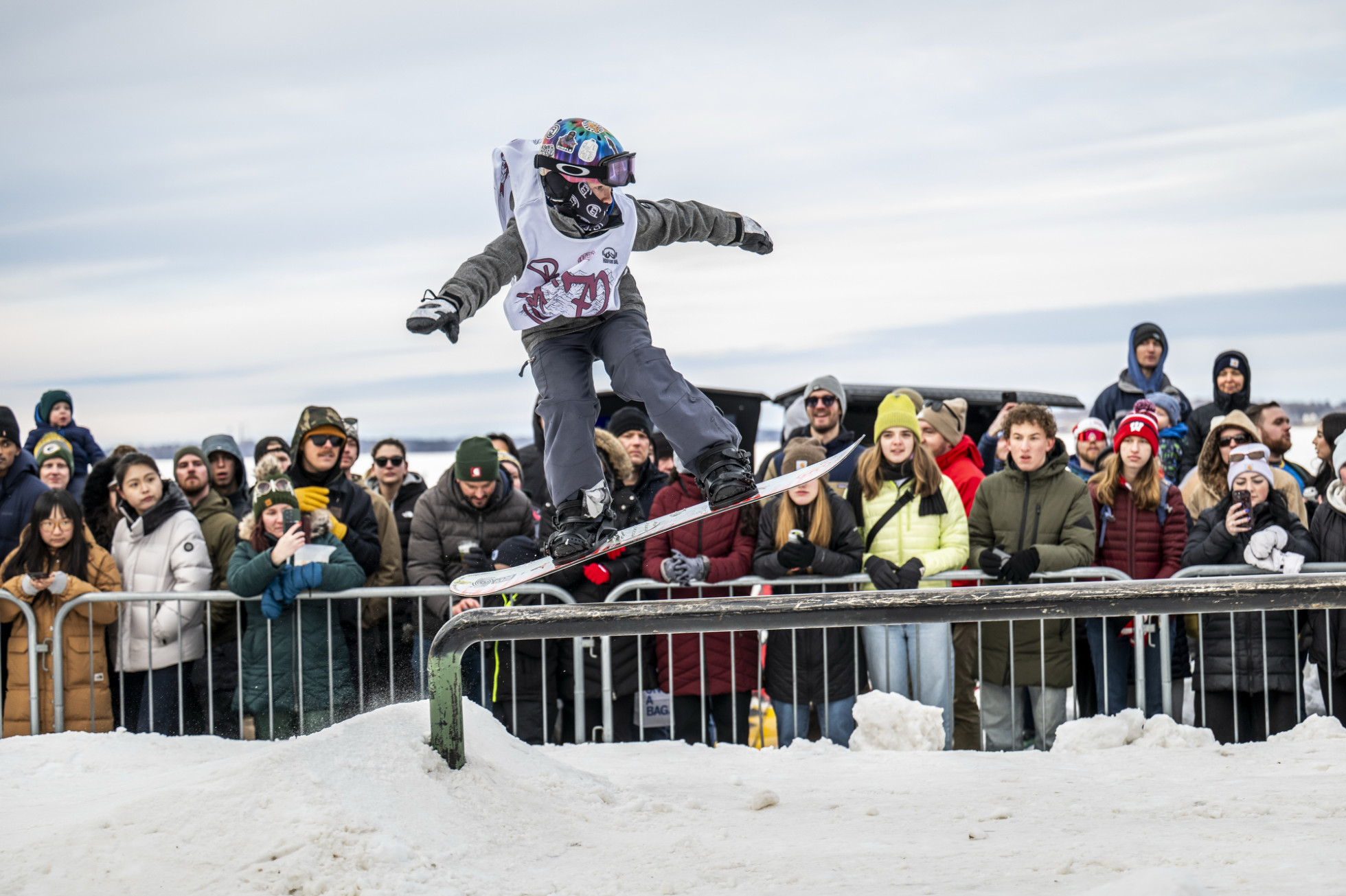 Spectators stand behind a metal gate as a person flies above a rail on their snowboard.
