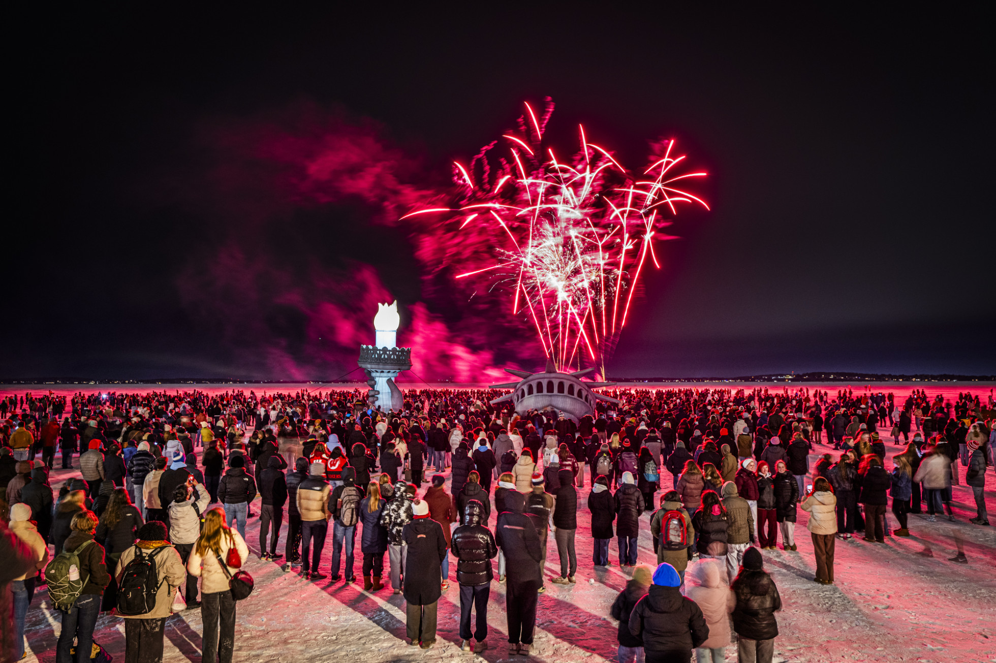 Hundreds of people standing on frozen Lake Mendota watch as fireworks launch into the night sky above an inflatable replica of the Statue of Liberty’s head, arm, and torch.