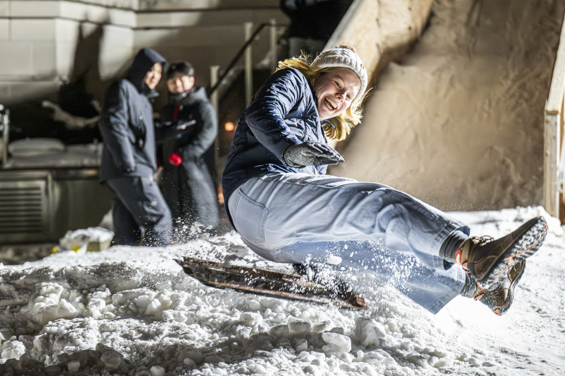 A blond woman smiles as she sleds down a ramp on a dining tray, her hair flying in the wind.