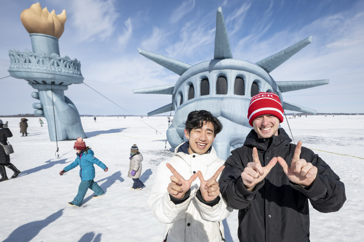 Two men smile and hold up Ws with their fingers with a partially submerged Statue of Liberty in the background. 