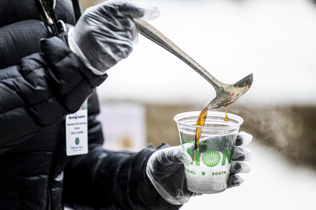 A person's arms pour boiling maple syrup from a ladle into a Union cup filled with clean snow, to make taffy.