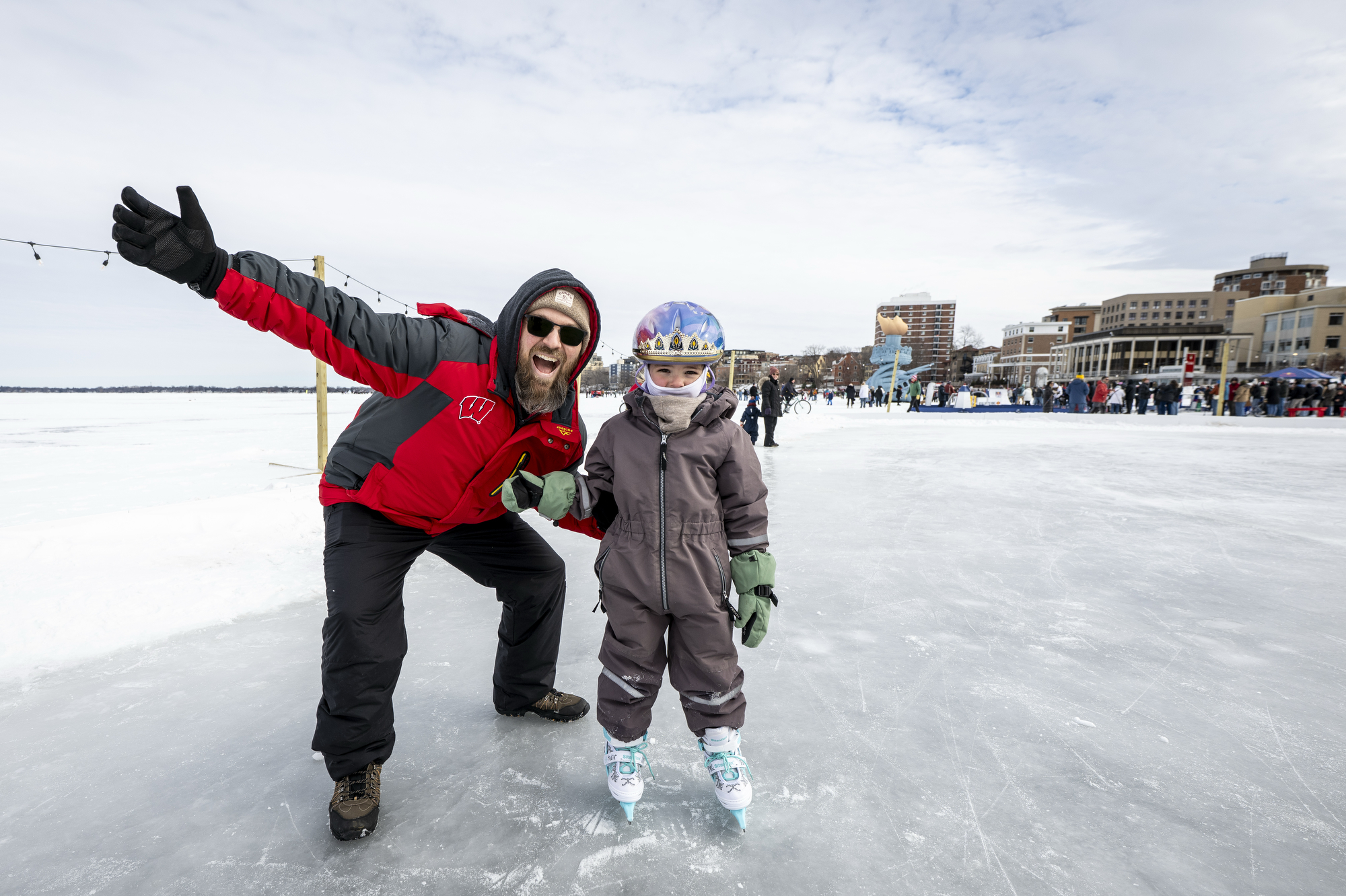 A man in Badger gear holds out his arms and smiles, crouching next to his 5-year-old daughter who is wearing a snowsuit and helmet.