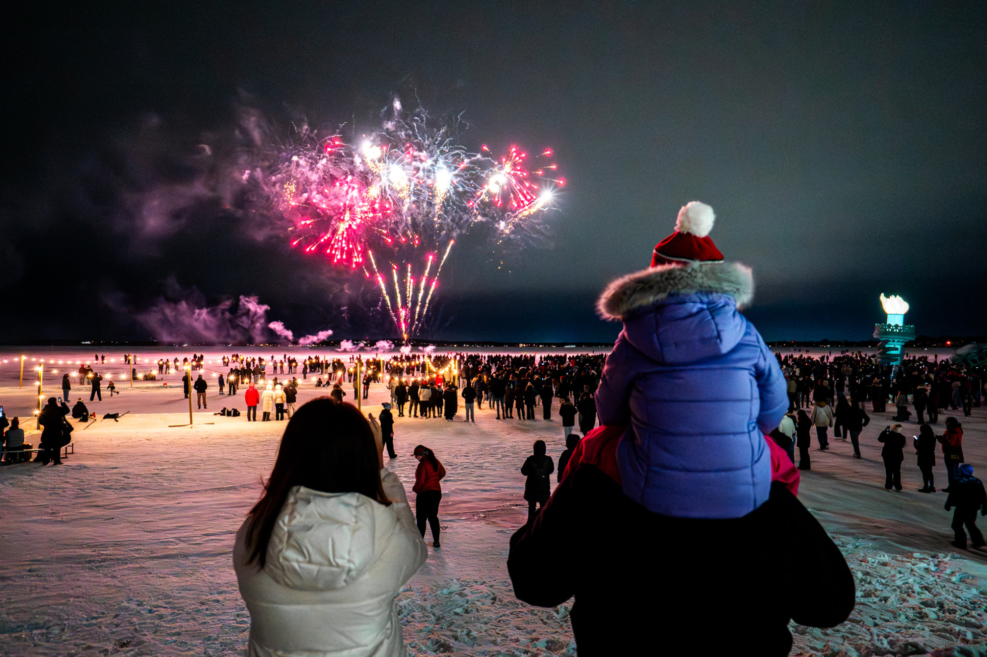 From behind, a child sits atop their parents shoulders while a crowd of hundreds gather on the frozen lake as red and white fireworks light up the night sky.