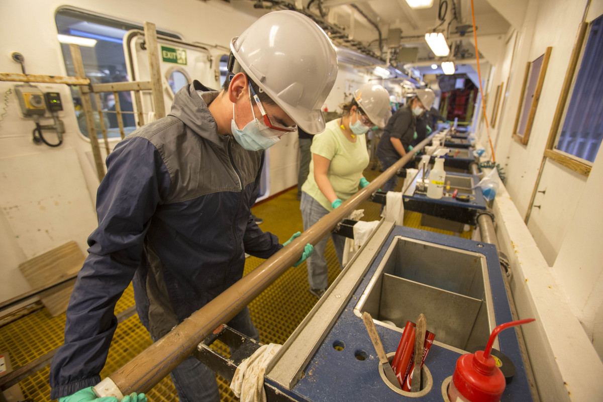 Lab specialists all wearing hard hats, face masks and latex glove hold a long metal pipe next to a series of utility sinks.