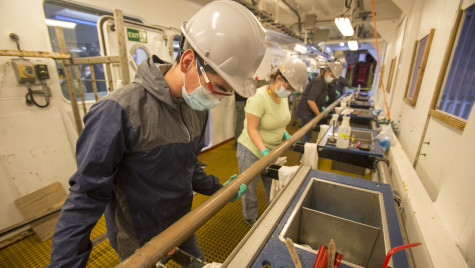 Lab specialists all wearing hard hats, face masks and latex glove hold a long metal pipe next to a series of utility sinks.