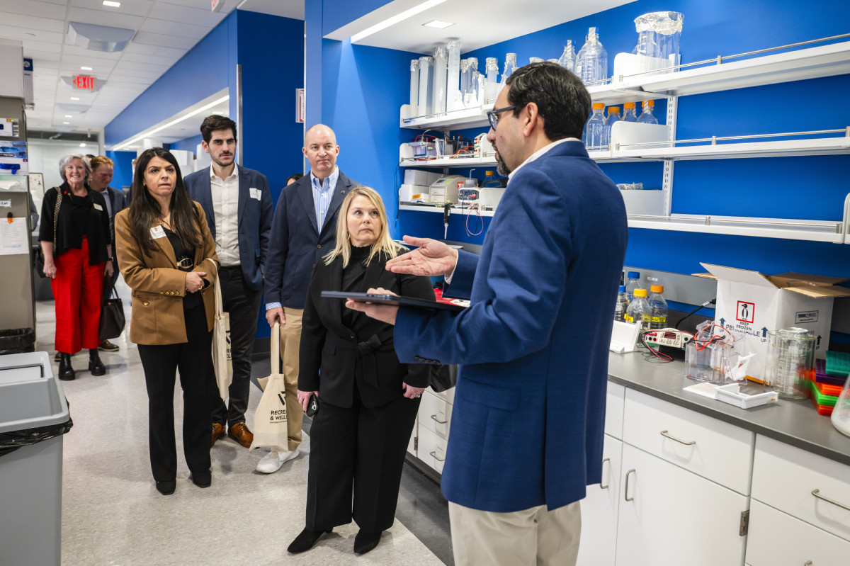 Visitors stand in a brightly lit laboratory while a staff member in a blue blazer speaks to the group and gestures toward the lab equipment on the counter.