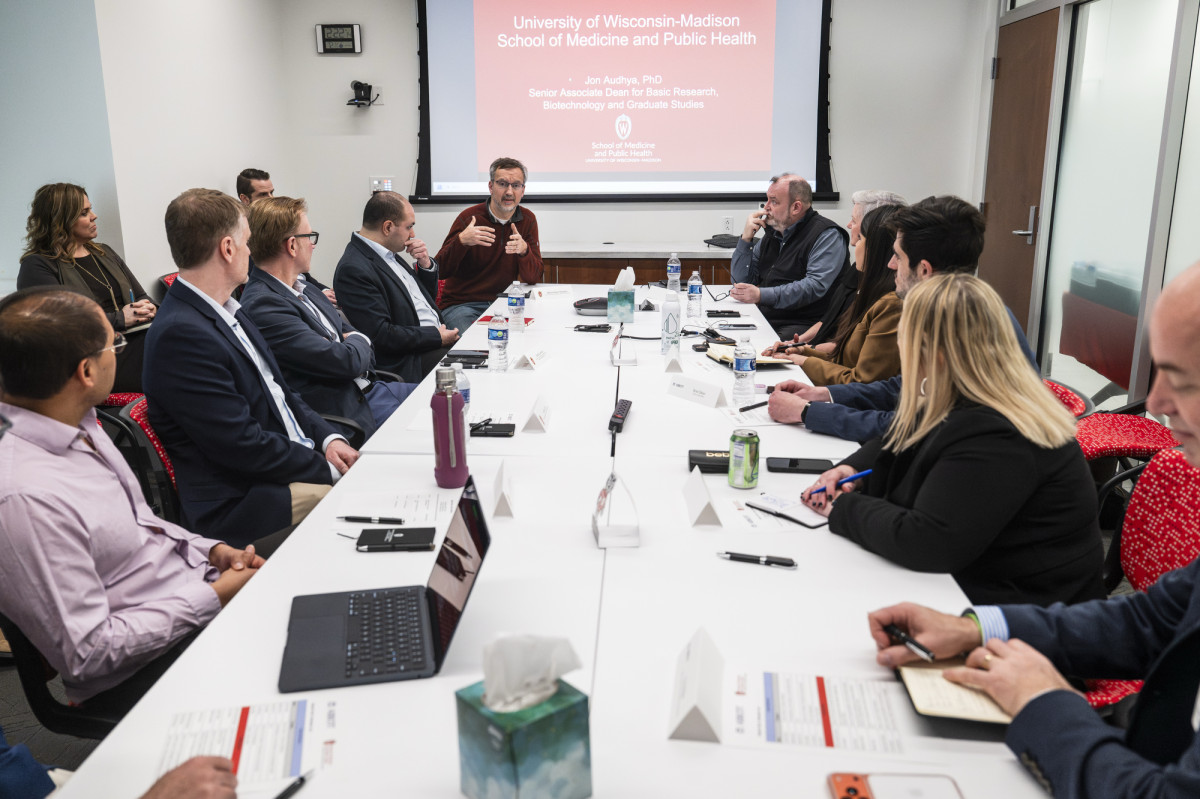 A group of people sit around a long conference table listening to a speaker during a meeting, with a presentation slide from the School of Medicine and Public Health displayed at the front of the room.