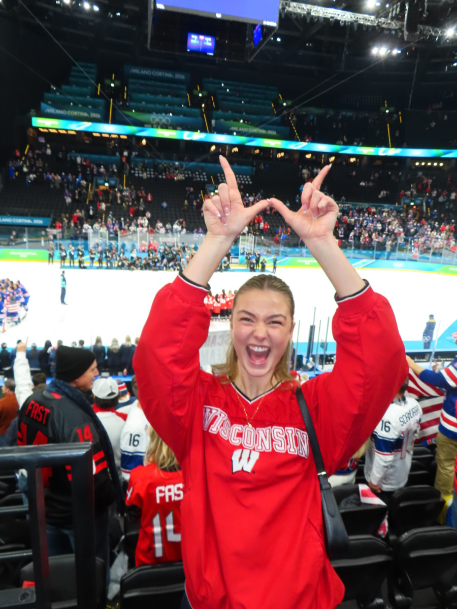 UW student Kate Sutton, wearing a red Badger sweater, makes a W with her hands in front of an Olympic hockey rink.