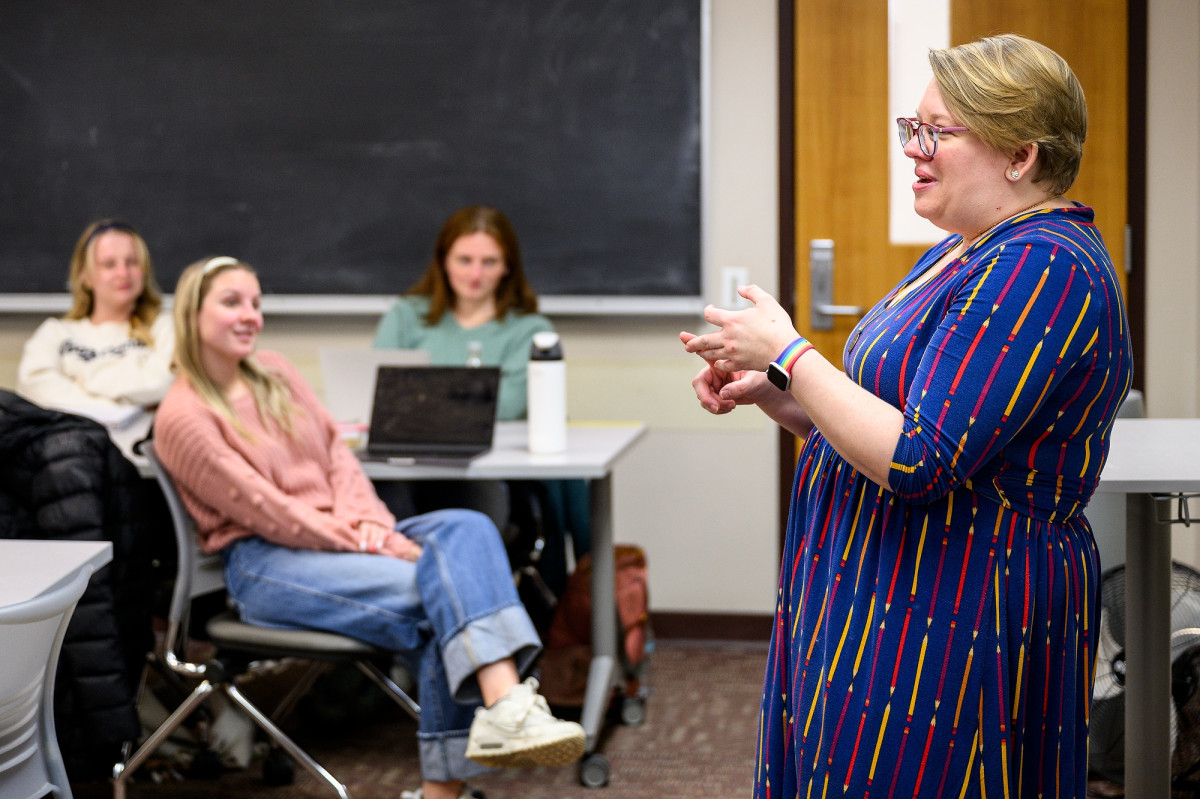 LB Klein stands in front of a classroom addressing students sitting at their grouped tables.