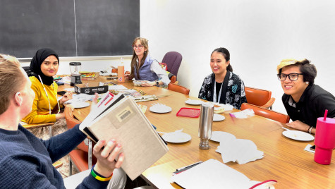 People, smiling and laughing, sit around a long table talking and looking at a stack of books.