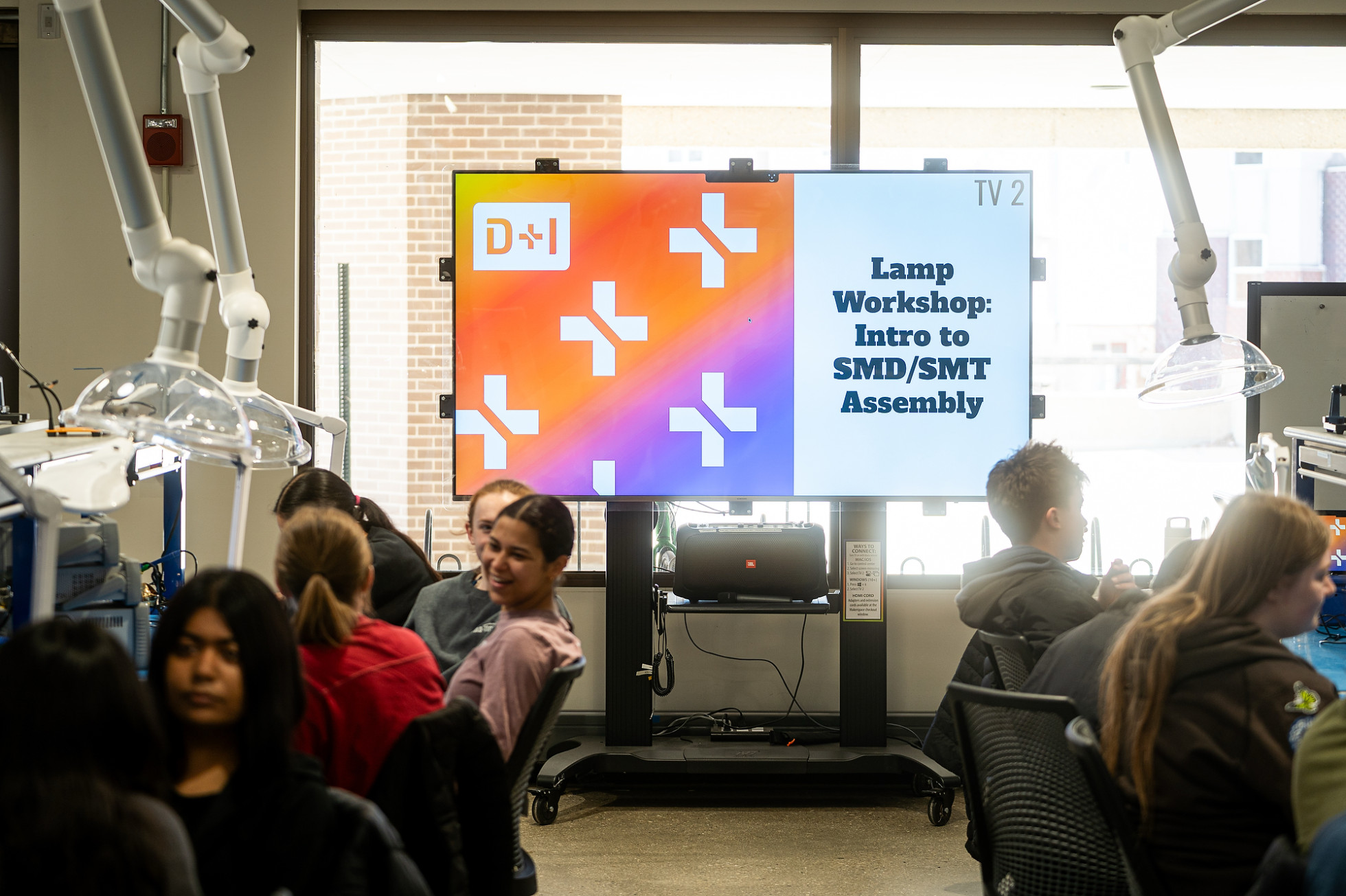 Two rows of students sitting at workstations prepare for a workshop, with a screen in the back that reads Lamp Workshop: Intro to SMD/SMT Assembly