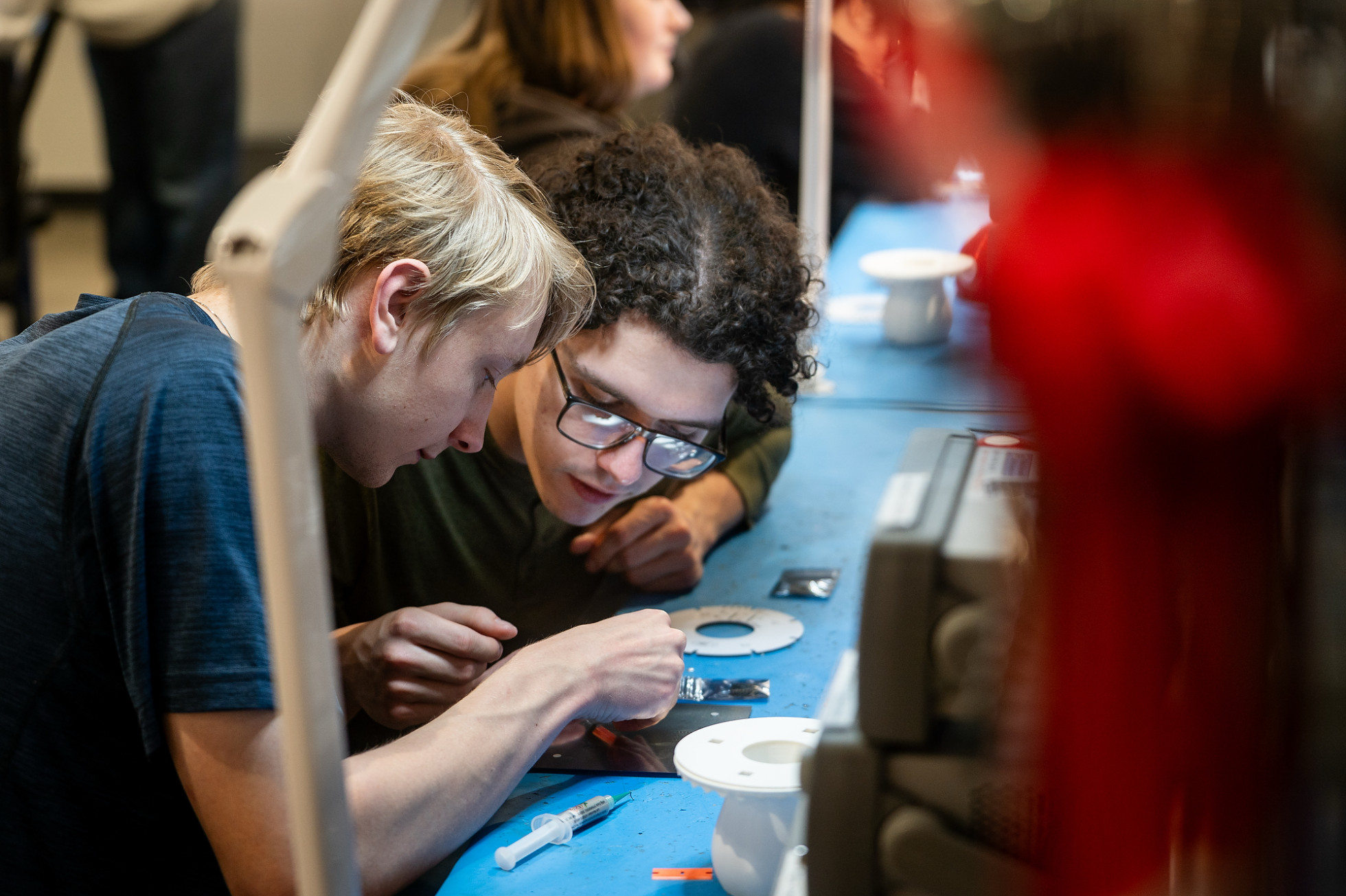 Two students stare closely at a circuit board at a workstation.