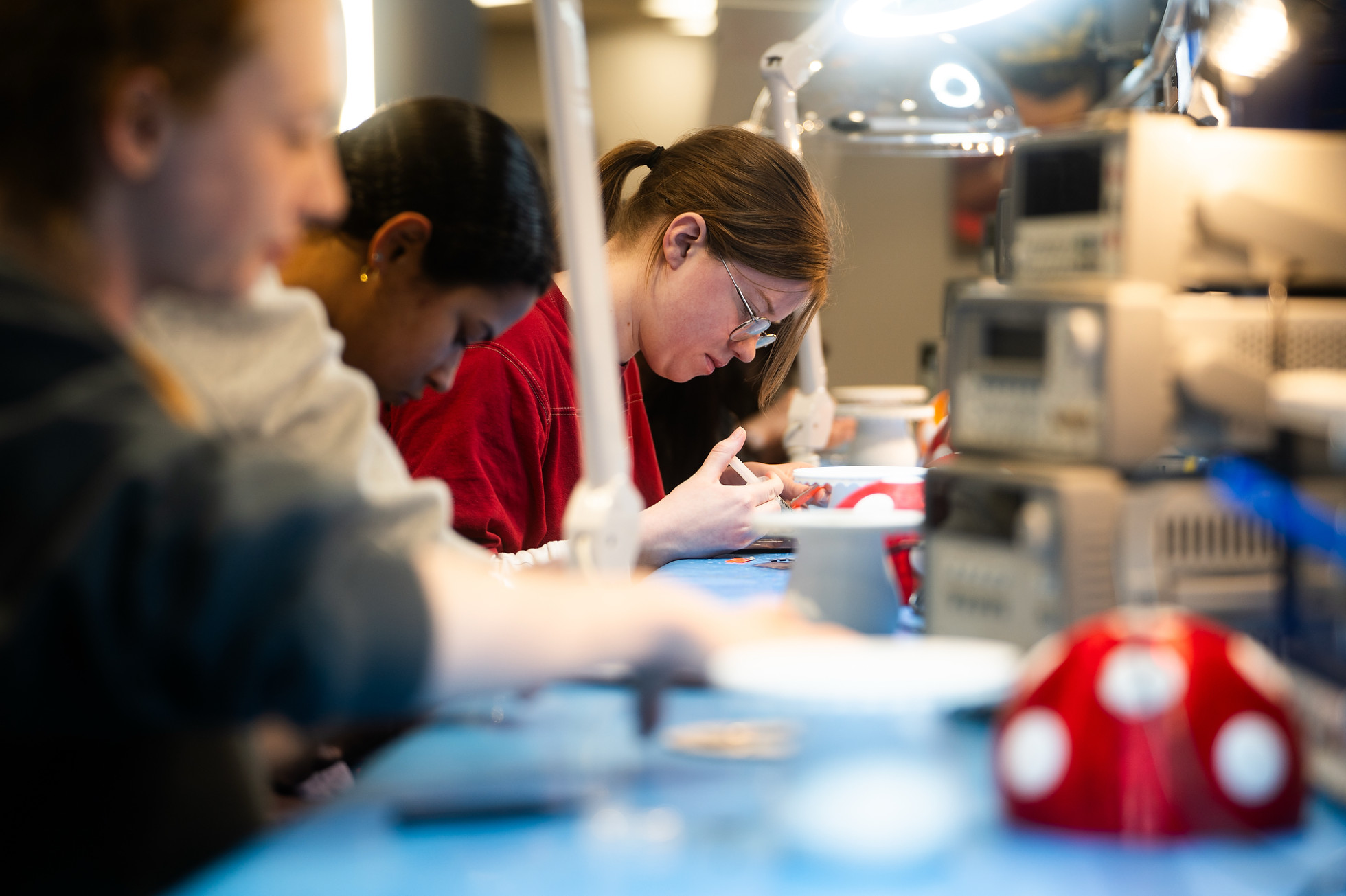 Three students at a blue table look focused while working on electronic parts. 