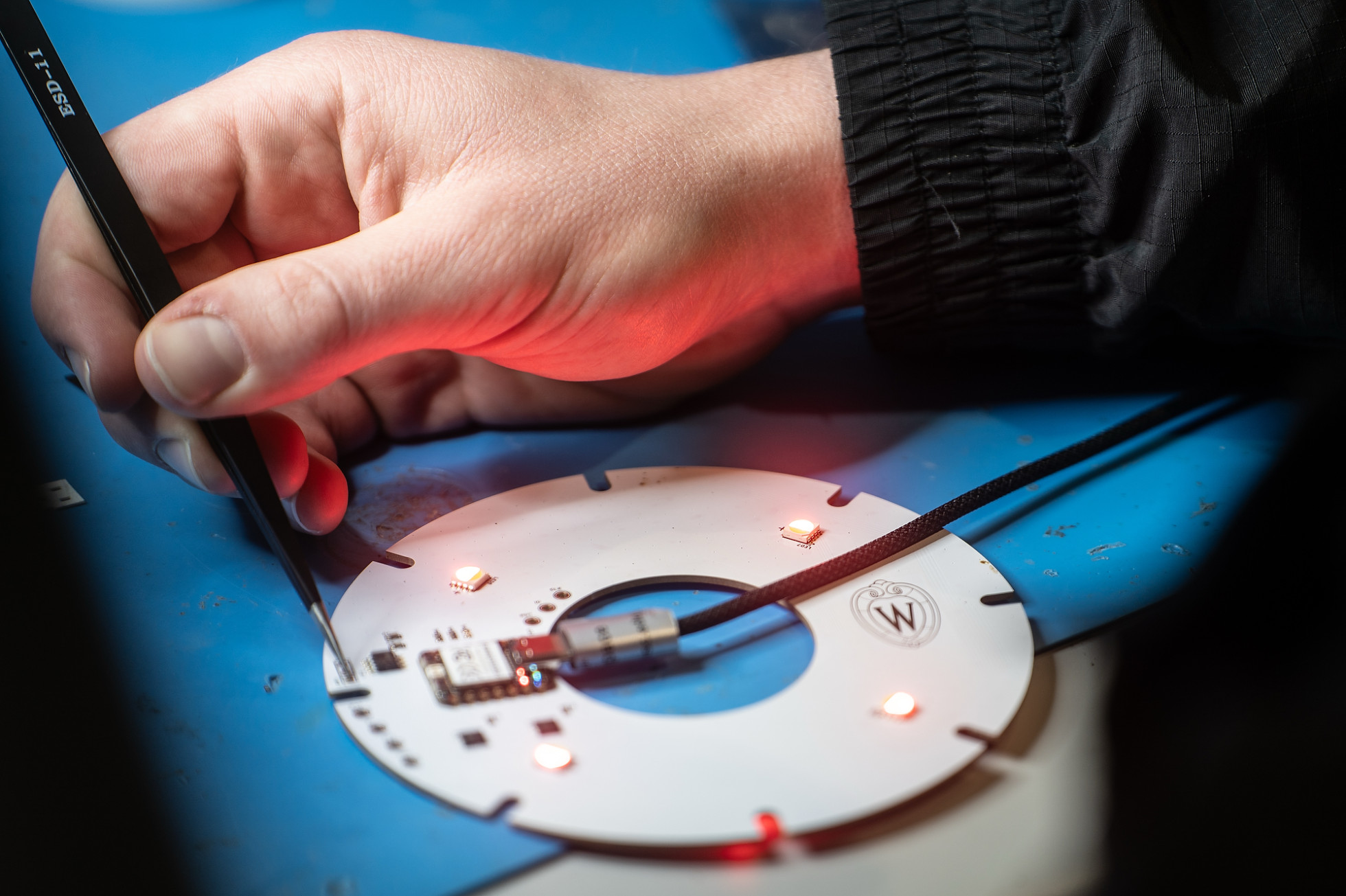 A participant's hand uses a tweezers to pick up a component on a circuit board.