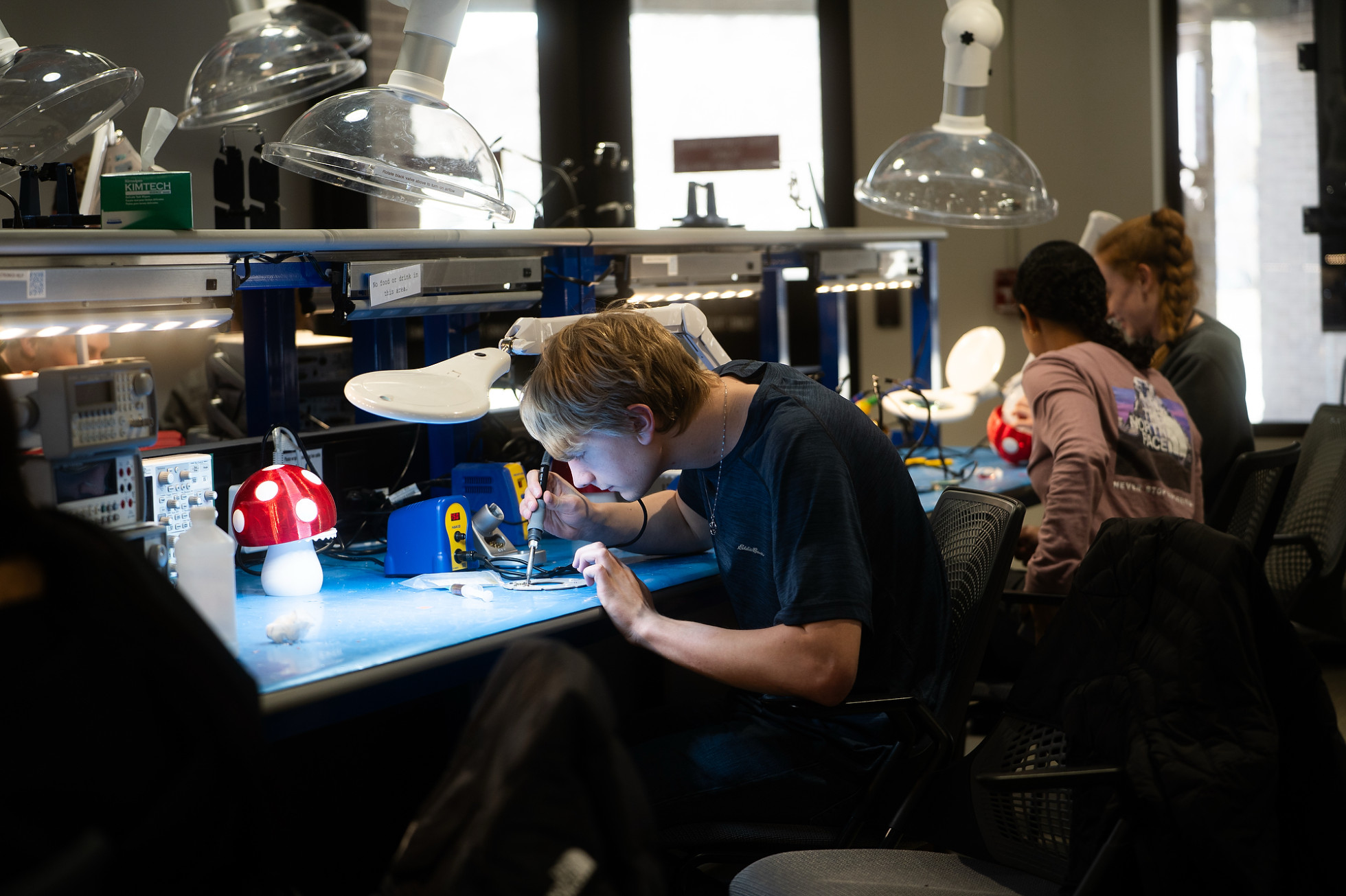 A student solders a circuit board at a workstation, with a small mushroom-style lamp nearby.  