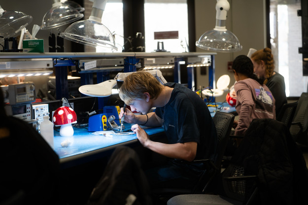 A student solders a circuit board at a workstation, with a small mushroom-style lamp nearby.