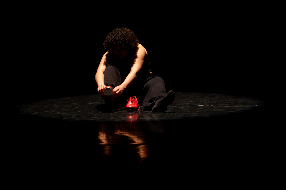 A student puts on red tap shoes and is illuminated on a dark stage by a single spotlight.