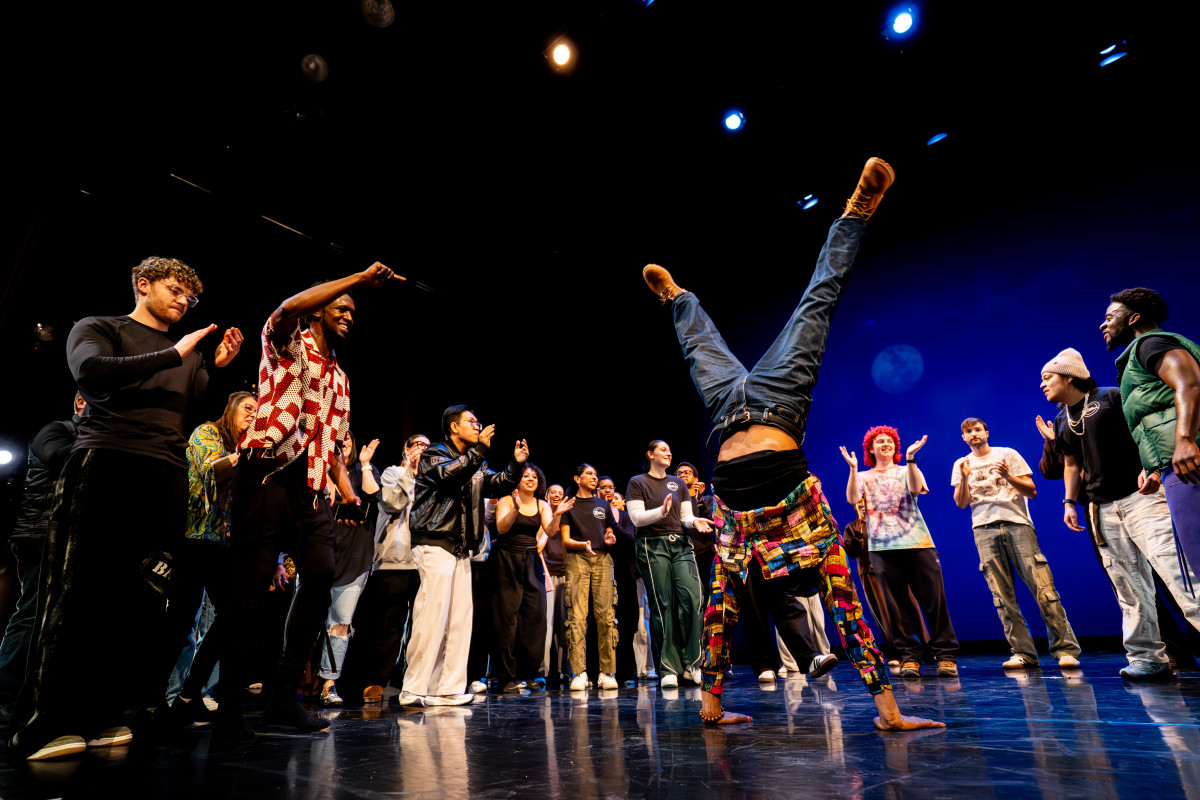 A person freestyle dances on stage surrounded by a group of people cheering them on.