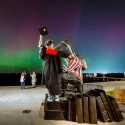A student wearing a graduation gown stands next to a Bucky Badger statue while holding his cap to the sky. In front of him, Northern Lights dance across the sky.